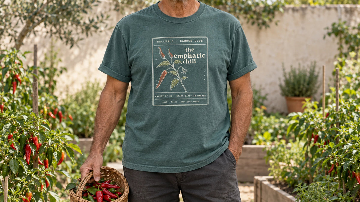 A man in the Blue Spruce Emphatic Chili tee stands by chili plants with glossy red and green peppers, holding a small wicker basket of red chili peppers.