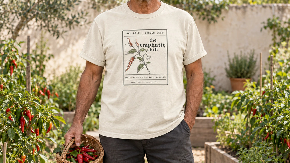 A man in the Ivory Emphatic Chili tee stands by chili plants with glossy red and green peppers, holding a small wicker basket of red chili peppers.