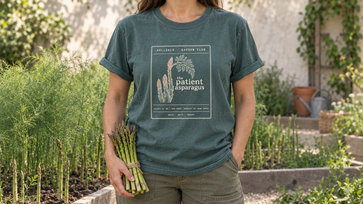 A woman in the Blue Spruce Patient Asparagus tee stands by an asparagus bed with tall ferny fronds behind young spears, holding a bundle of fresh green asparagus spears tied with twine.