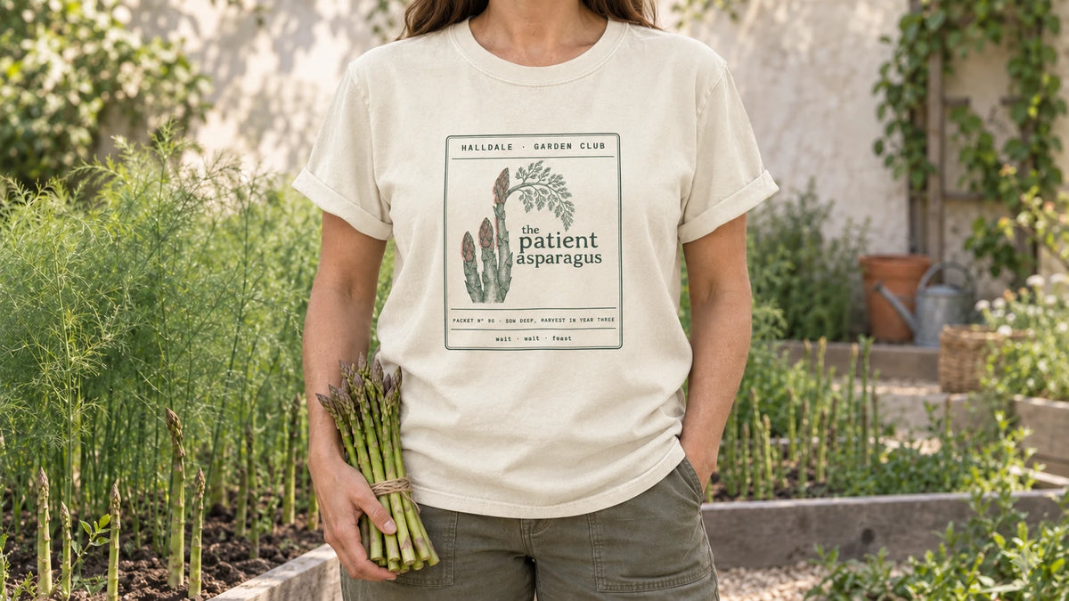 A woman in the Ivory Patient Asparagus tee stands by an asparagus bed with tall ferny fronds behind young spears, holding a bundle of fresh green asparagus spears tied with twine.