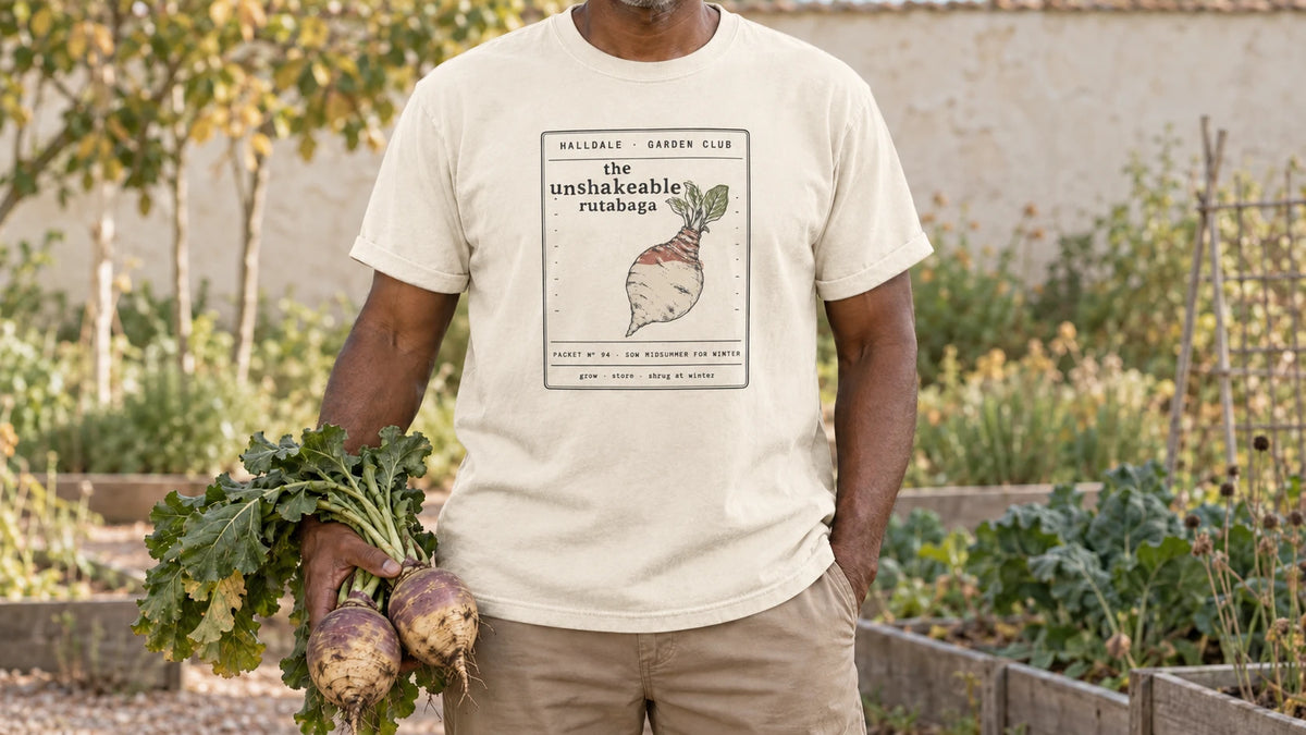 A man in the Ivory Unshakeable Rutabaga tee stands by a late fall root bed, holding two purple-shouldered rutabagas with their greens.