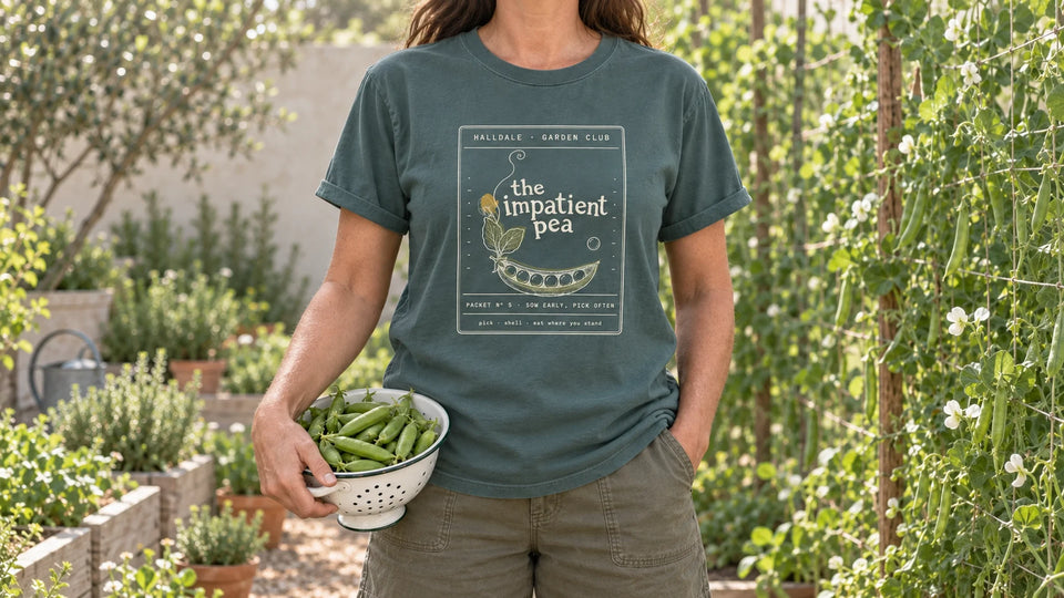 A woman in the Blue Spruce Impatient Pea tee stands beside a pea trellis, holding a colander of fresh pea pods at her hip.